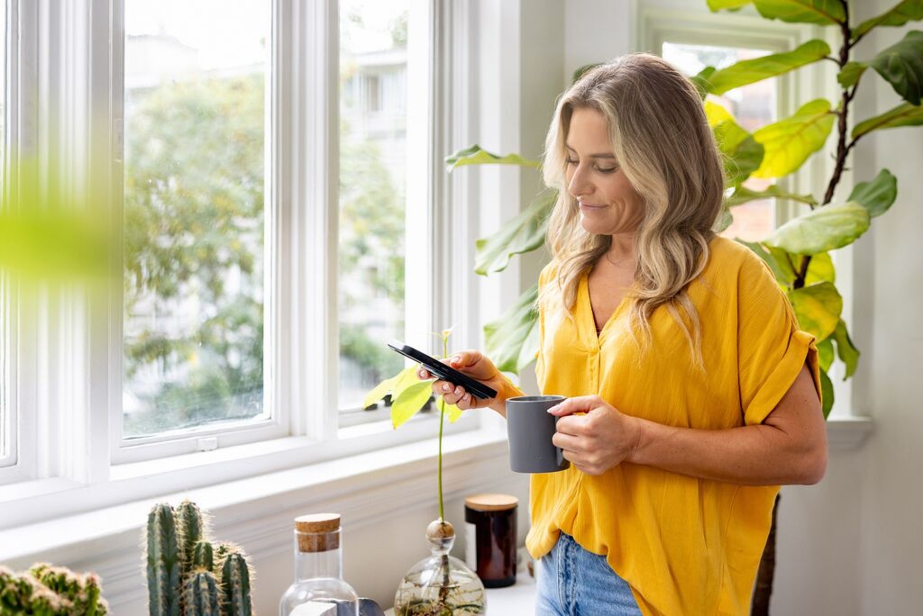 Een vrouw staat met een kop koffie bij haar raam en kijkt naar haar telefoon